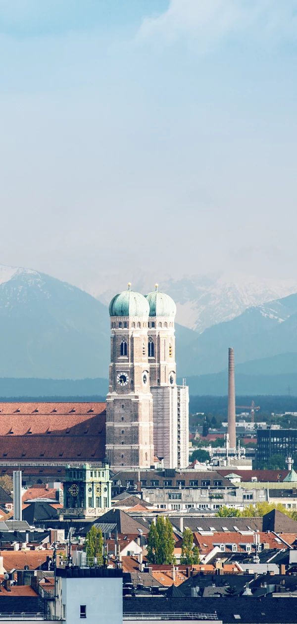 Blick auf die Skyline von München mit historischen Gebäuden wie der Frauenkirche und dem Alten Peter, im Hintergrund sind die schneebedeckten Alpen zu sehen.