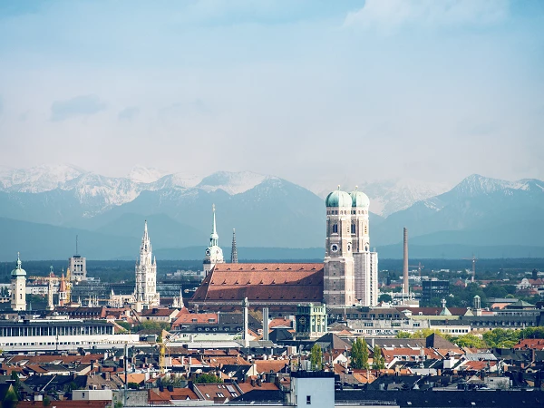 Blick auf die Skyline von München mit historischen Gebäuden wie der Frauenkirche und dem Alten Peter, im Hintergrund sind die schneebedeckten Alpen zu sehen.
