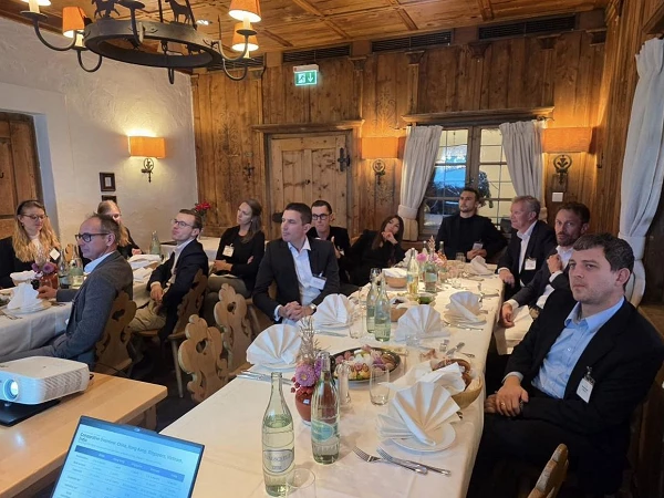 Eine Gruppe von zehn bis fünfzehn Personen sitzt an einem festlich gedeckten Tisch in einem rustikalen Holzraum, während sie einem Vortrag auf einer Leinwand oder einem Projektor zuhören. Einige Teilnehmer machen Notizen, und auf dem Tisch stehen Wasserflaschen und dekorative Blumenarrangements.