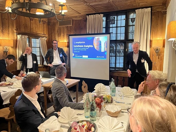 Eine Gruppe von Menschen sitzt an einem Tisch in einem rustikalen Raum, während ein Präsentator neben einem Bildschirm steht, auf dem Informationen zu einem "Lunchon Insights" Event angezeigt werden. Am Tisch sind Speisen und Getränke sichtbar, und die Teilnehmer scheinen aktiv an der Diskussion teilzunehmen.