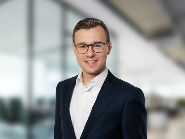 Ein junger Mann in einem schwarzen Anzug und einer weißen Hemd steht lächelnd vor einem modernen, hellen Büro mit verschwommenem Hintergrund.