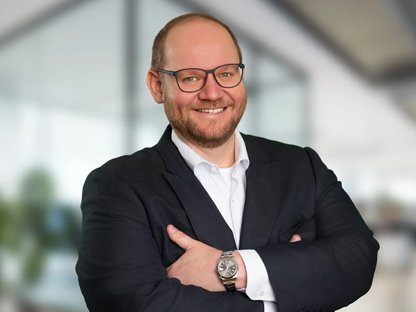 Ein lächelnder Mann mit Brille steht in einem modernen Büro. Er trägt einen dunkelblauen Anzug und hat die Arme vor der Brust verschränkt. Im Hintergrund sind unscharfe Büroelemente zu sehen.