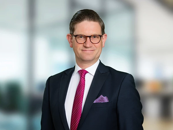 Ein Mann in einem dunkelblauen Anzug mit einem weißen Hemd und einer pinken Krawatte lächelt in die Kamera. Er hat eine Brille auf und eine lila gestreifte Einstecktuch in der Brusttasche. Im Hintergrund sind unscharfe Büroelemente zu erkennen.