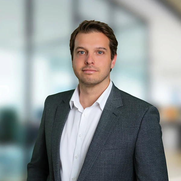 Ein Mann in einem grauen Anzug steht vor einem unscharfen Bürohintergrund und schaut direkt in die Kamera.
