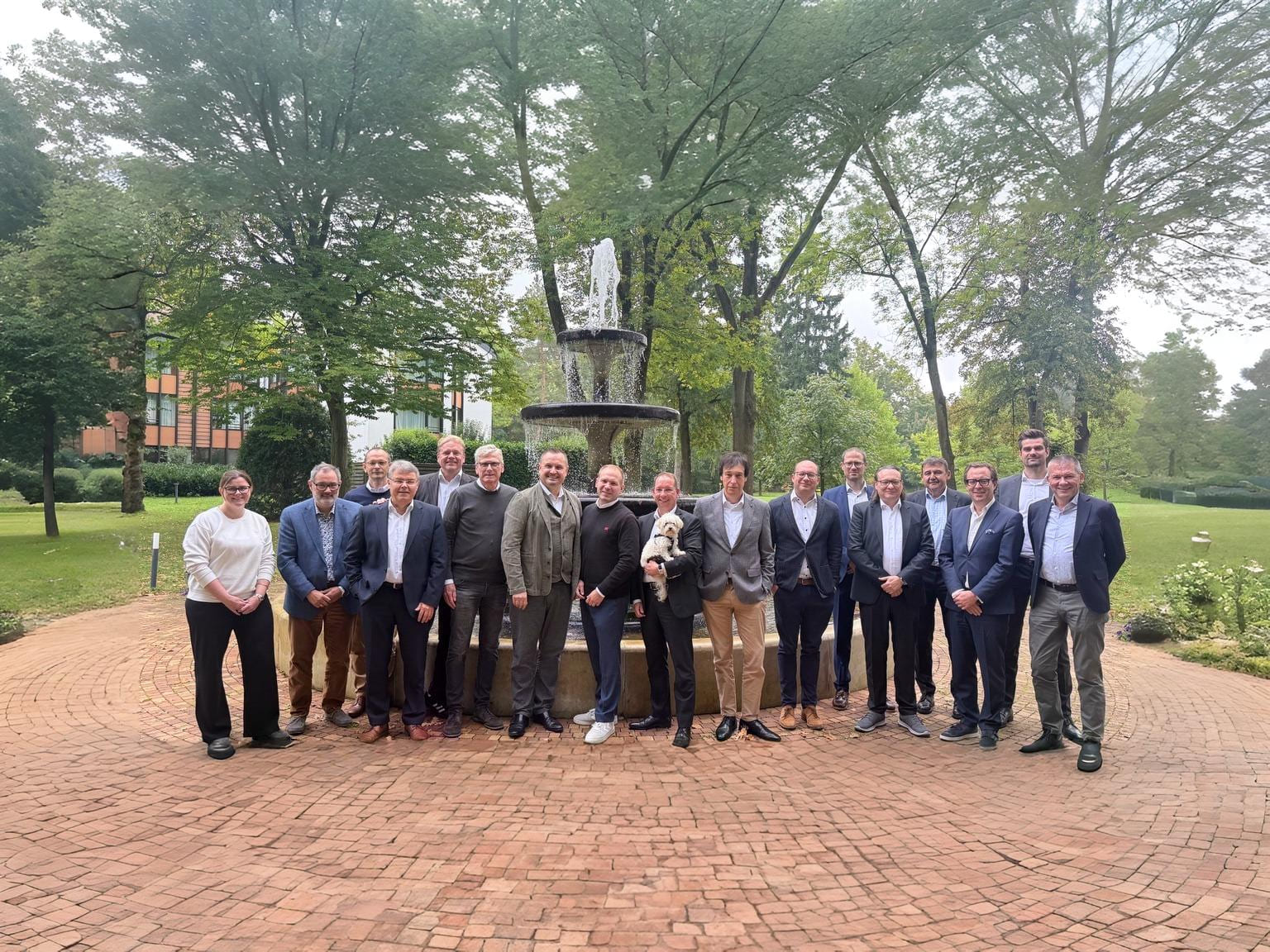 Eine Gruppe von 21 Personen posiert lächelnd vor einem Springbrunnen in einem parkähnlichen Garten. Bäume und grüne Wiesen sind im Hintergrund sichtbar.