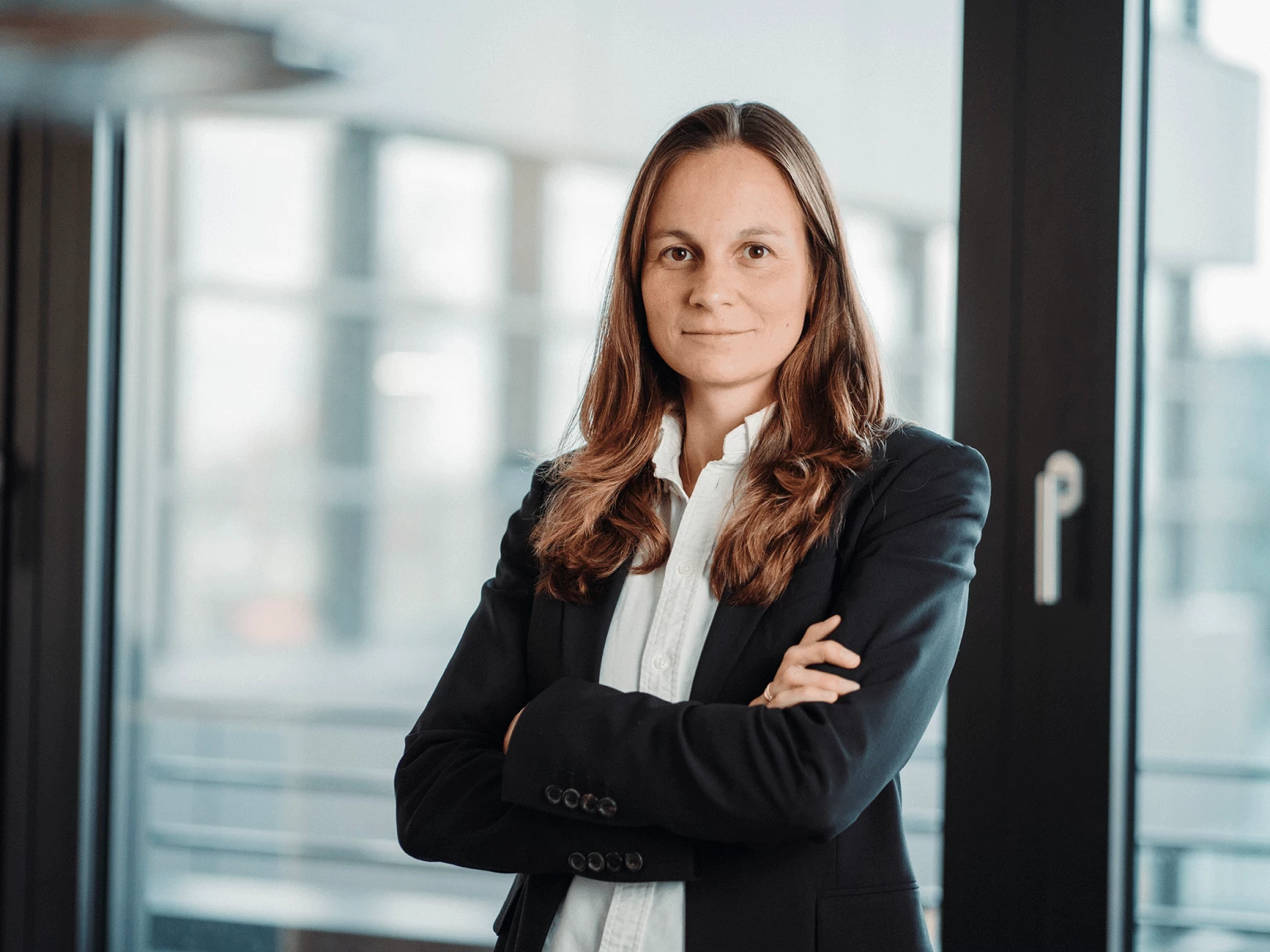 Eine Frau mit langen, braunen Haaren steht mit verschränkten Armen in einem modernen Büro vor einem großen Fenster und schaut direkt in die Kamera. Sie trägt einen schwarzen Blazer und ein weißes Hemd.