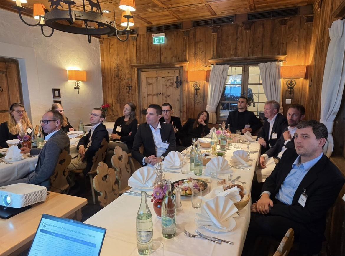 Eine Gruppe von zehn bis fünfzehn Personen sitzt an einem festlich gedeckten Tisch in einem rustikalen Holzraum, während sie einem Vortrag auf einer Leinwand oder einem Projektor zuhören. Einige Teilnehmer machen Notizen, und auf dem Tisch stehen Wasserflaschen und dekorative Blumenarrangements.