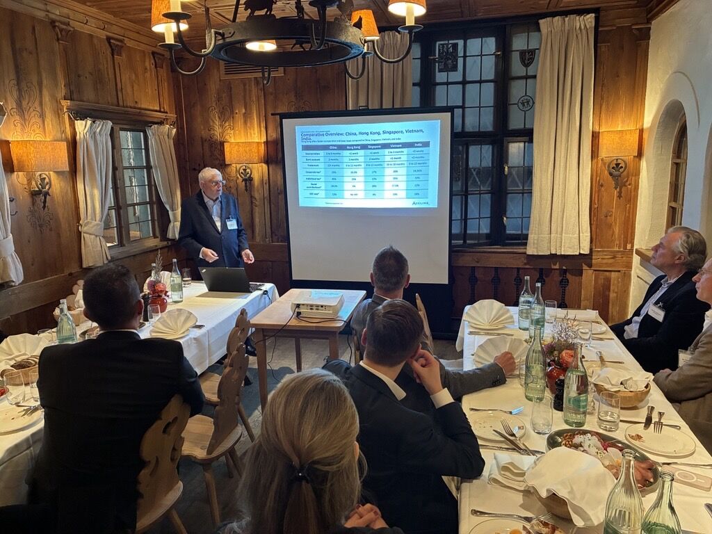 Ein Mann steht vor einem Bildschirm in einem rustikalen Raum und präsentiert eine Tabelle mit Daten. Im Vordergrund sitzen Personen an einem gedeckten Tisch, einige hören aufmerksam zu.